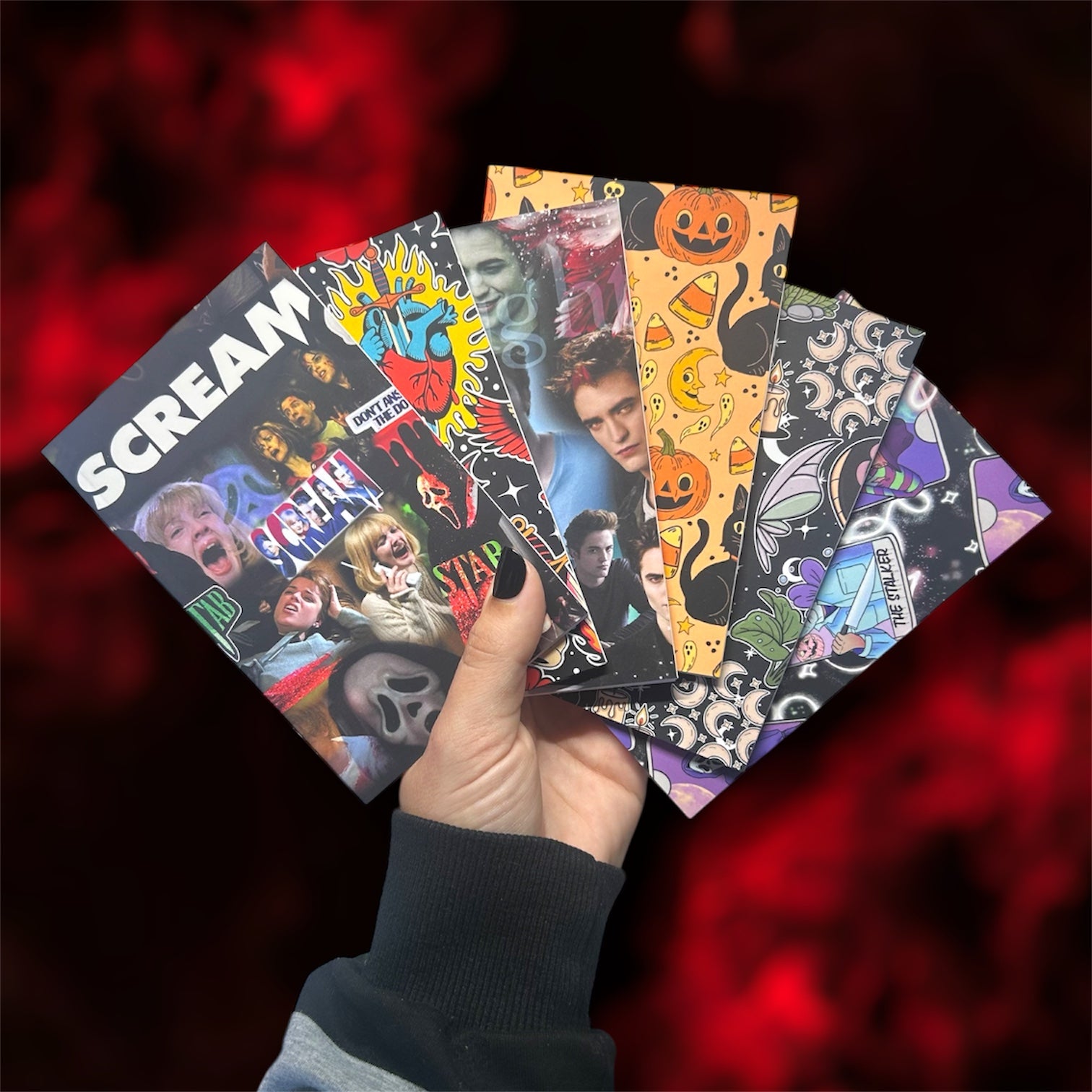 Hand holding several pocket notebooks, including Scream, traditional tattoo art, Twilight, Halloween, cottagecore, and horror tarot, on a blurred blue background