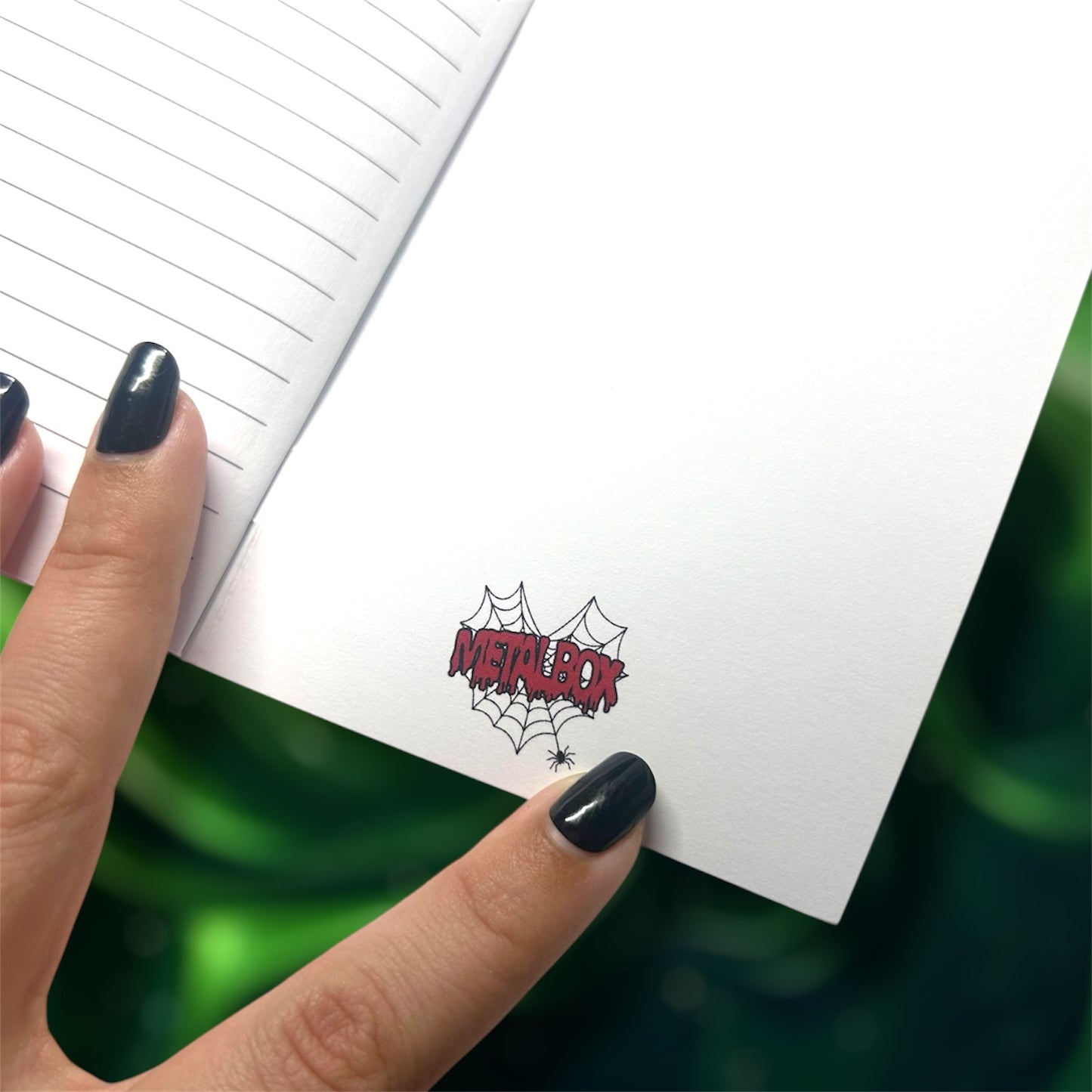 Hand holding an open pocket notebook with the Metalbox logo on the inside back cover on a blurred green background