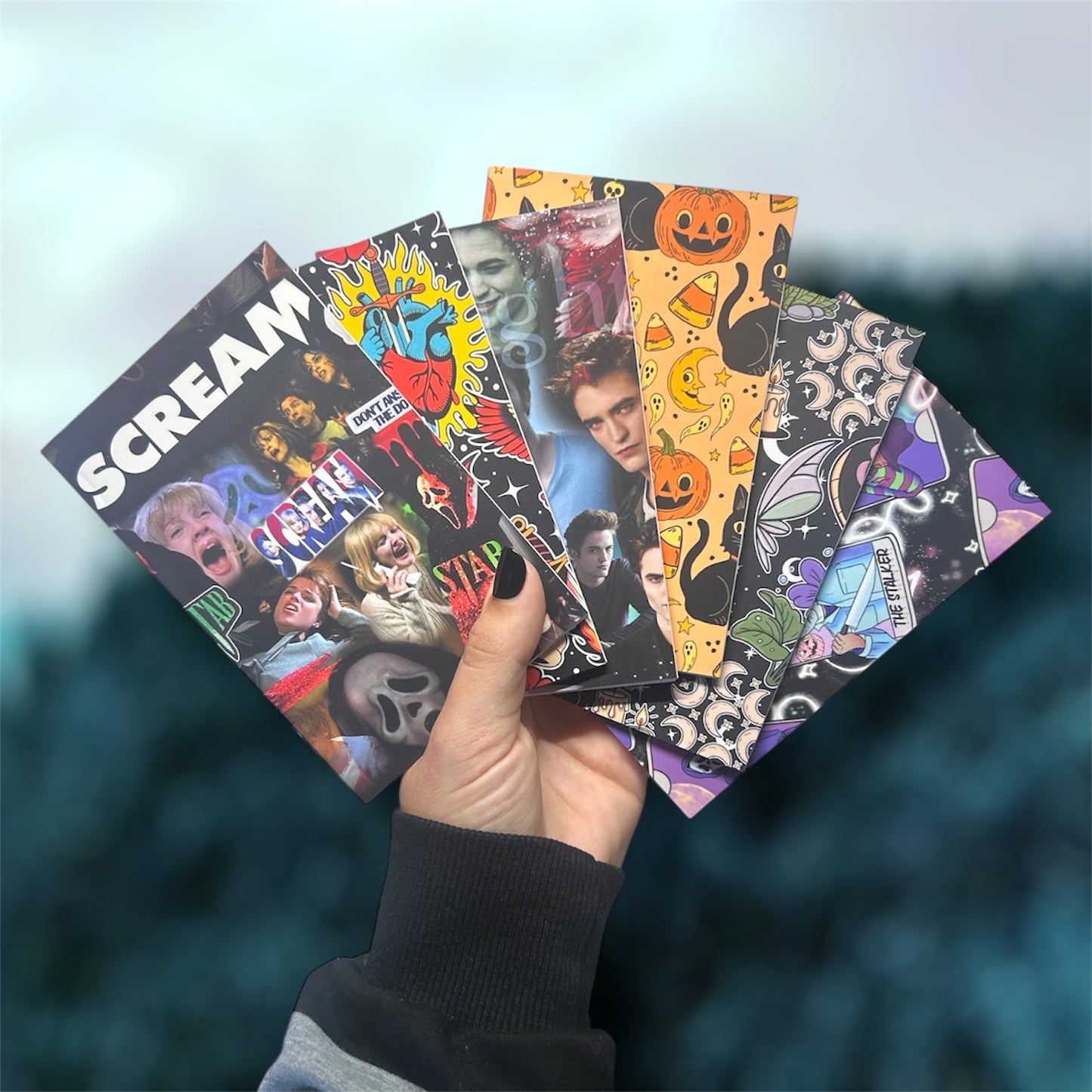 Hand holding several pocket notebooks, including Scream, traditional tattoo art, Twilight, Halloween, cottagecore, and horror tarot, on a blurred blue background