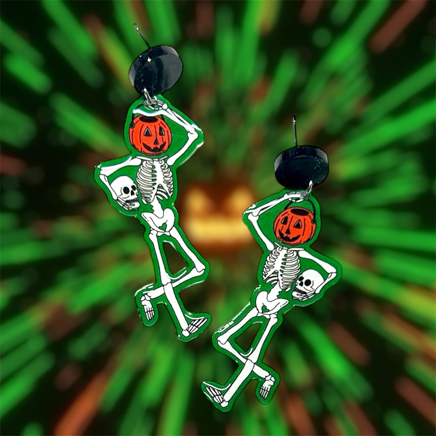 Glowing green trick-or-treat skeleton earrings with a Halloween pumpkin bucket head holding his skull on a blurred green background.