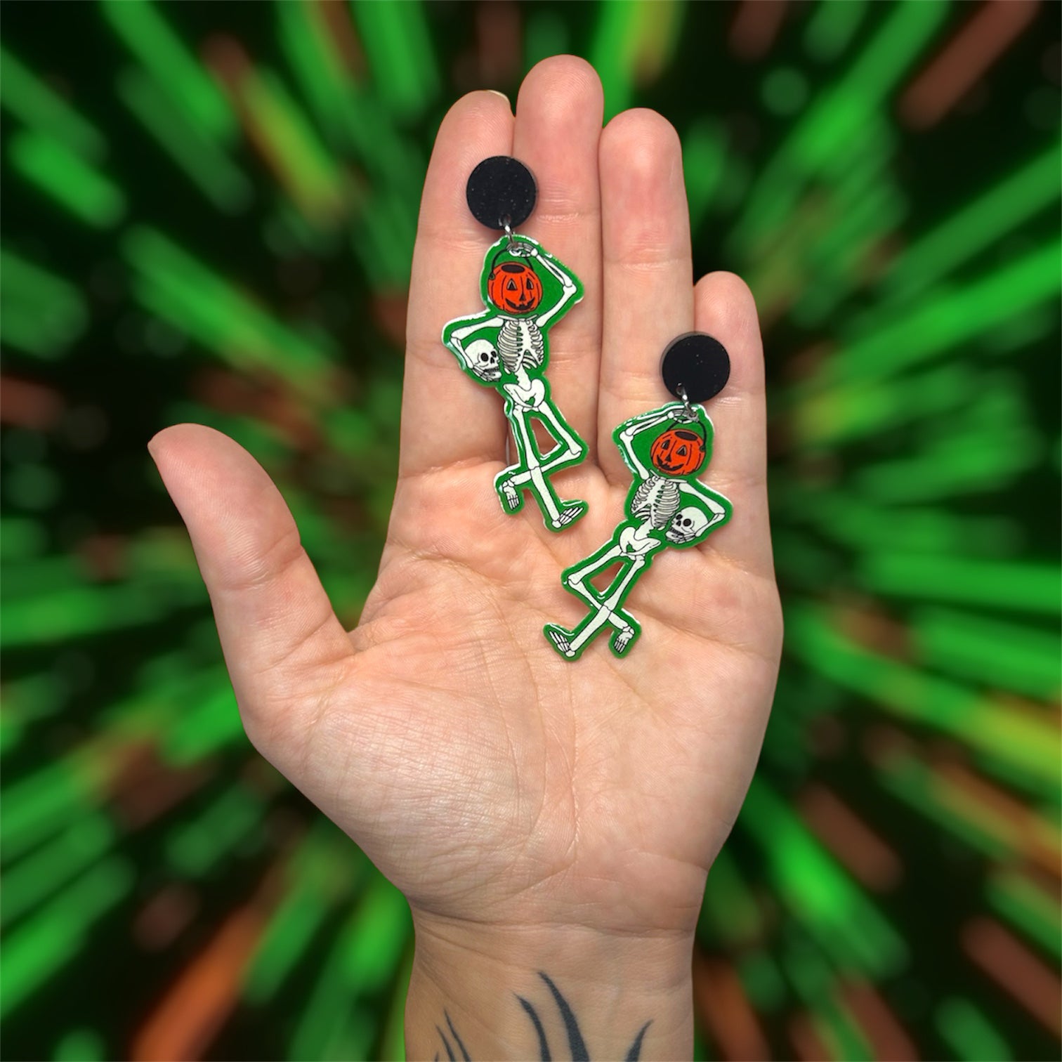 Hand holding glowing green trick-or-treat skeleton earrings with a Halloween pumpkin bucket head holding his skull on a blurred green background.