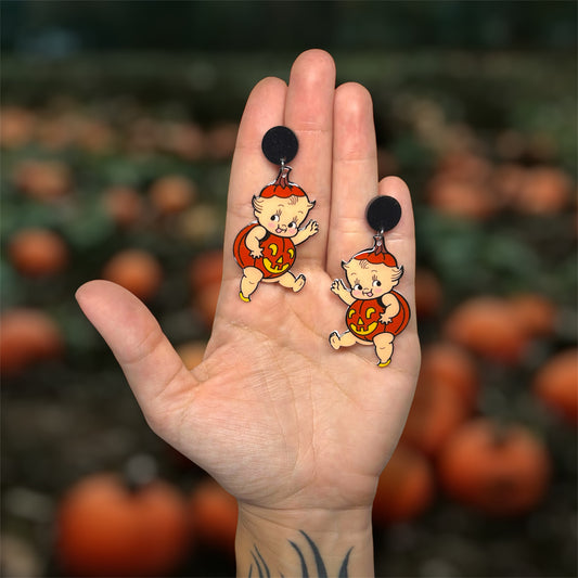 Hand holding Halloween cherub baby kewpie pumpkin jack-o-lantern earrings on a blurred pumpkin patch background.