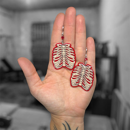 Hand holding glowing red ribcage beaded earrings on a blurred black and white background.