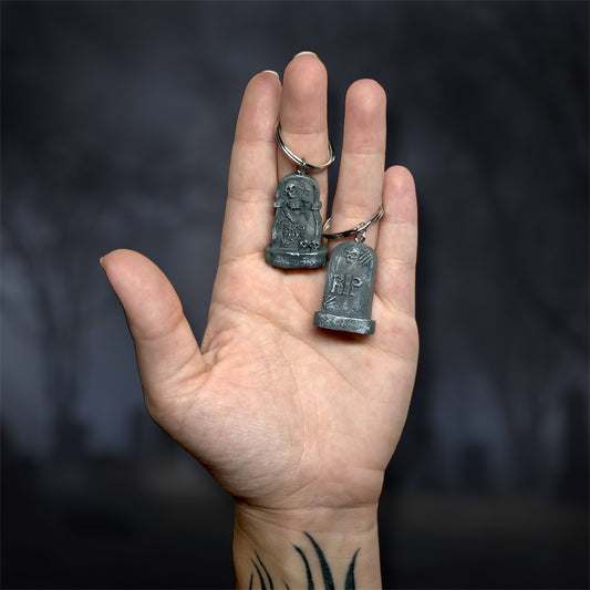 Hand holding two tombstone keychains with 'RIP' on a dark background