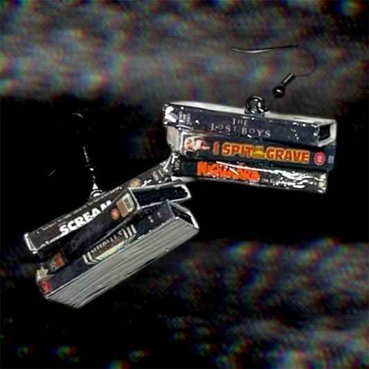 A pair of stacked VHS tape statement earrings, featuring the movies Scream, Chopping Mall, Halloween 3, The Lost Boys, I Spit on Your Grave, and A Nightmare on Elm Street, shown against a dark, grainy TV static background.