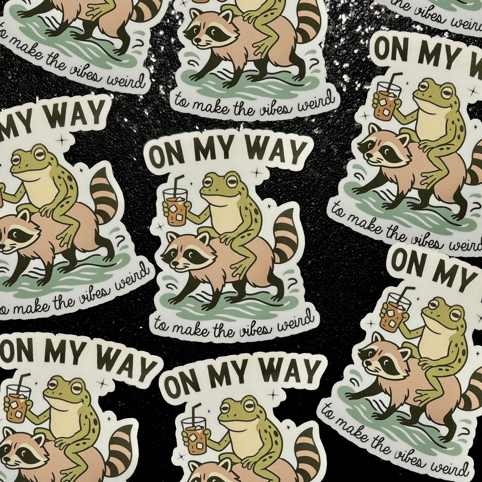 Stickers of a frog drinking iced coffee riding a raccoon with text 'On my Way to Make the Vibes Weird' on a black background