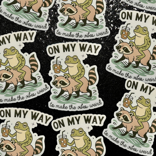 Stickers of a frog drinking iced coffee riding a raccoon with text 'On my Way to Make the Vibes Weird' on a black background
