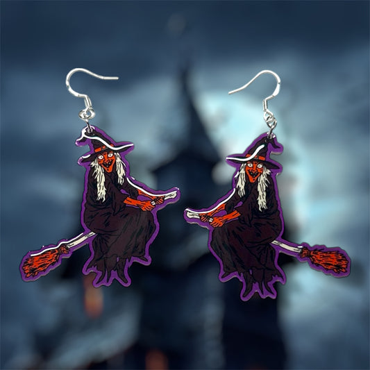 Halloween earrings of a witch with red skin riding a broom and wearing a black cloak and a black witch hat on a blurred background.