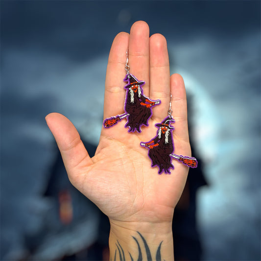 Hand holding Halloween earrings of a witch with red skin riding a broom and wearing a black cloak and a black witch hat on a blurred background.