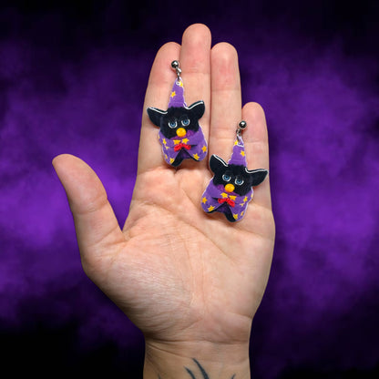 Hand holding earrings of a classic Furby dressed as a wizard with a wizard hat and cape on a smoky purple background.