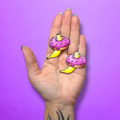 Hand holding a pair of bananas inside pink sprinkle donuts resin dangle earrings on a purple background
