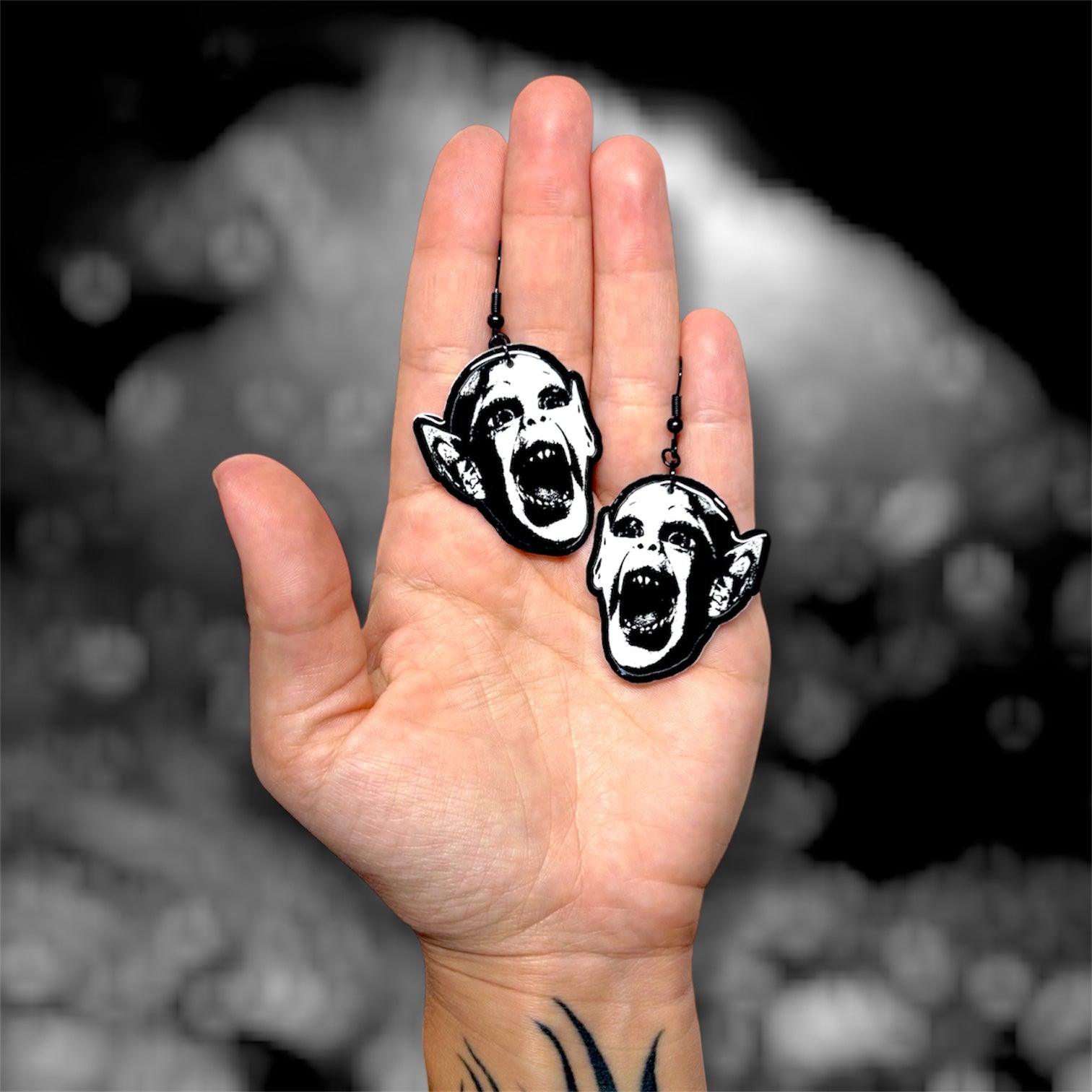 Hand holding a pair of bat boy spooky resin dangle earrings on a blurry black and white background