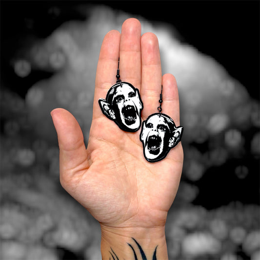 Hand holding a pair of bat boy spooky resin dangle earrings on a blurry black and white background