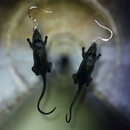 A pair of black rat dangle earrings on a blurred sewer background