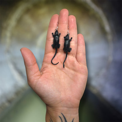 A hand holding a pair of black rat dangle earrings on a blurred sewer background