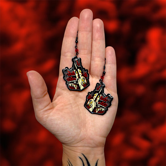 Hand holding a pair of burning church resin dangle earrings with red glass beads and black earring hooks on a blurry red background