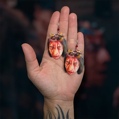 Hand holding a pair of Terrifier 2 Allie's mom bloody candy bowl head beaded resin statement earrings on a blurred background