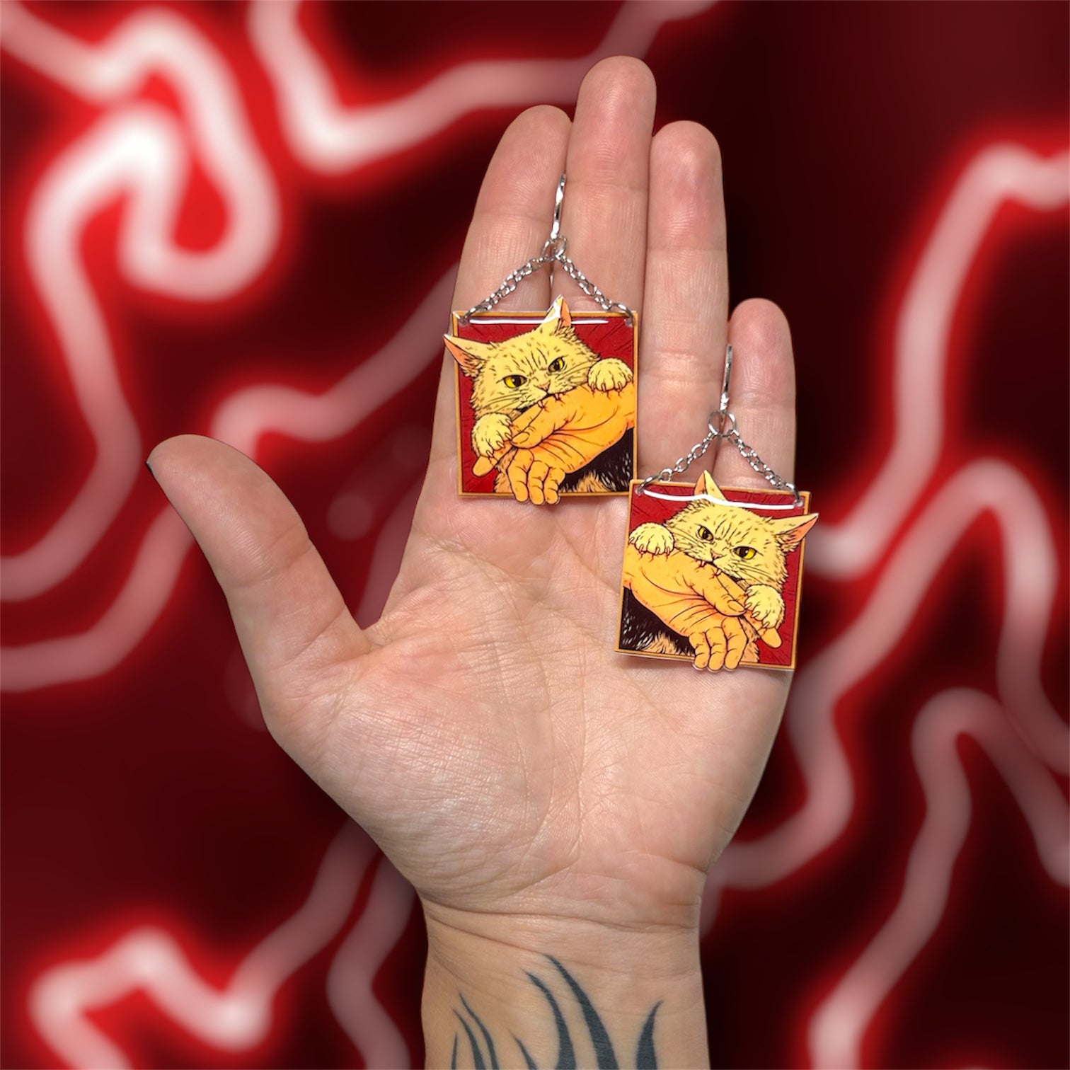Hand holding angry cat biting hand comic book panel art resin statement earrings on a blurry red background