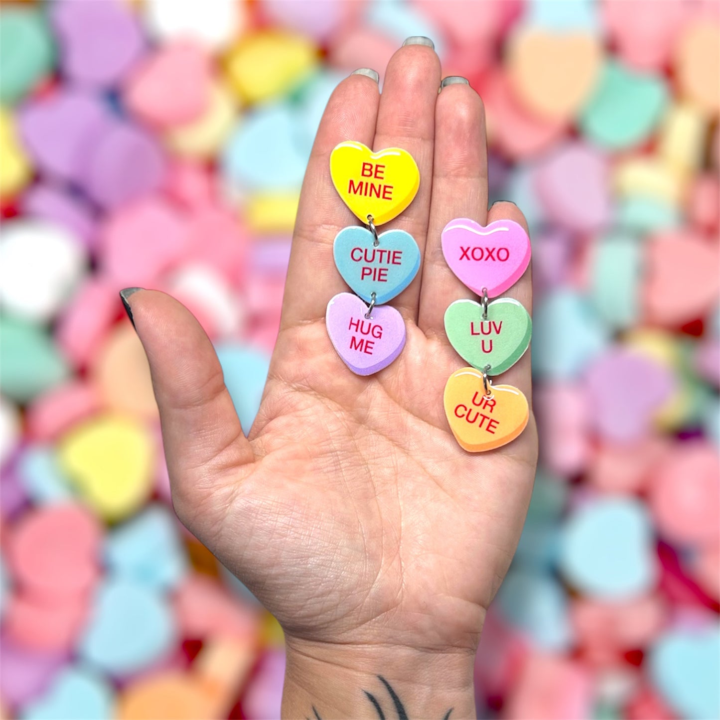 Hand holding a pair of Valentine's Day Sweethearts conversation hearts stacked dangle earrings on a blurred colorful background