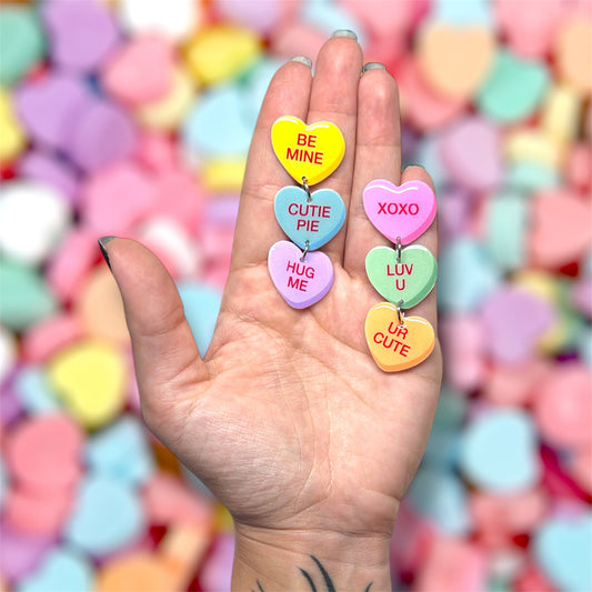 Hand holding a pair of Valentine's Day Sweethearts conversation hearts stacked dangle earrings on a blurred colorful background