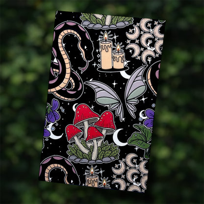Black pocket notebook with colorful mystical design featuring mushrooms, candles, a snake, and butterflies on a blurred green leafy background.