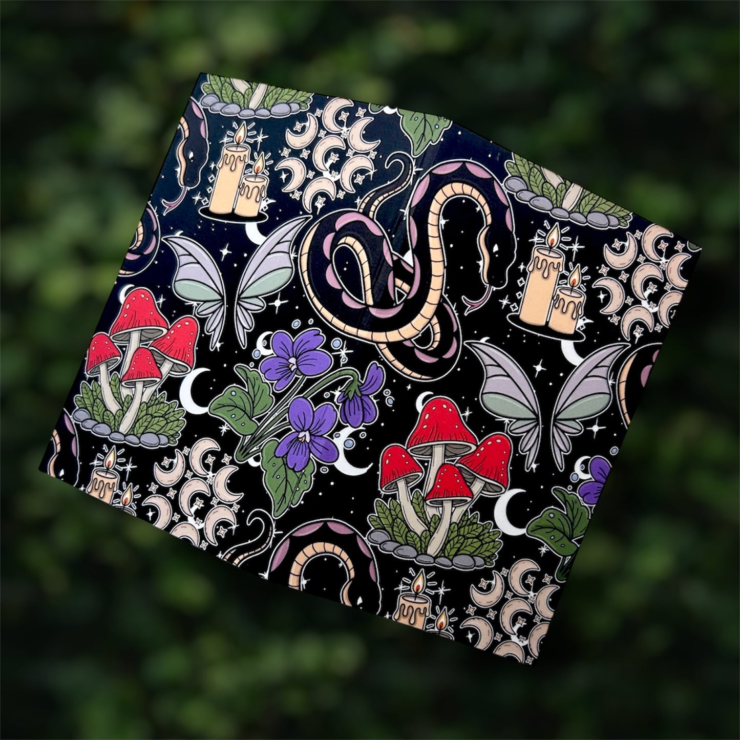 Open black pocket notebook with colorful mystical design featuring mushrooms, candles, a snake, and butterflies on a blurred green leafy background.