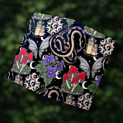 Open black pocket notebook with colorful mystical design featuring mushrooms, candles, a snake, and butterflies on a blurred green leafy background.