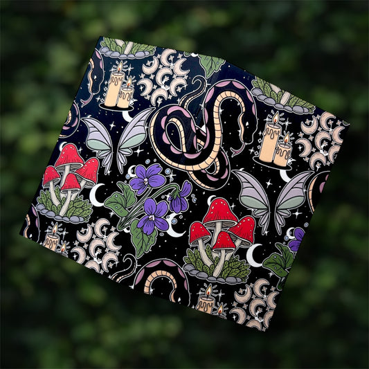 Open black pocket notebook with colorful mystical design featuring mushrooms, candles, a snake, and butterflies on a blurred green leafy background.