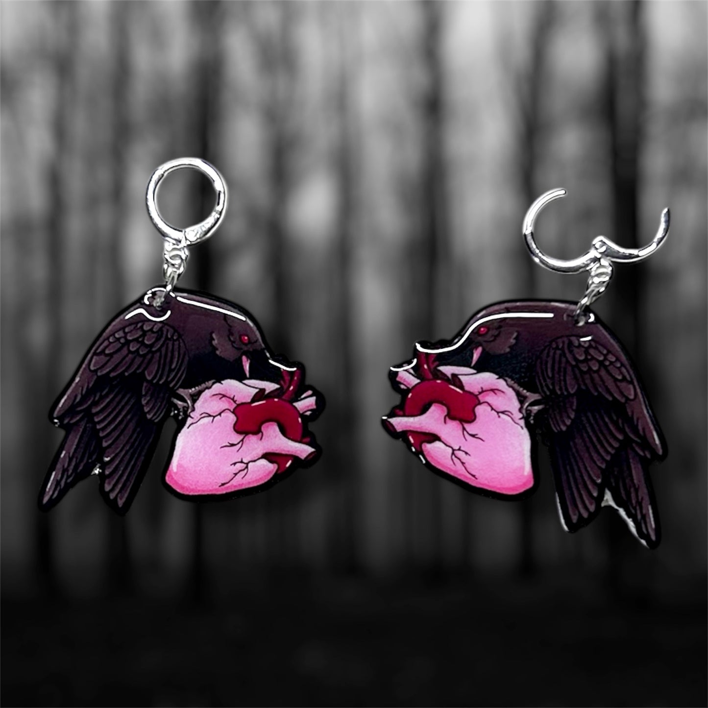 A pair of dangle earrings of a black crow with red eyes eating a heart on a blurred black and white background