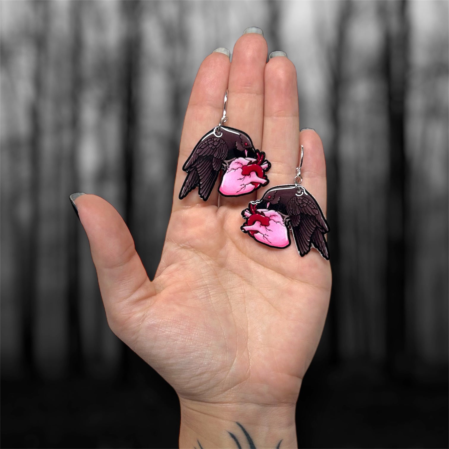 Hand holding a pair of dangle earrings of a black crow with red eyes eating a heart on a blurred black and white background