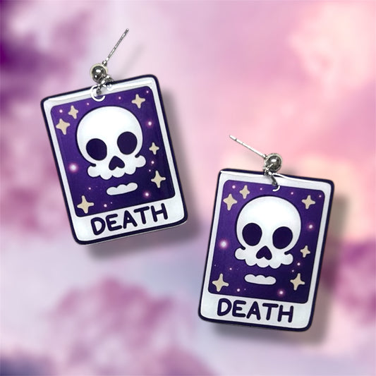 Sparkly skull Death tarot card witch resin dangle earrings on a blurry pink, purple, and white background