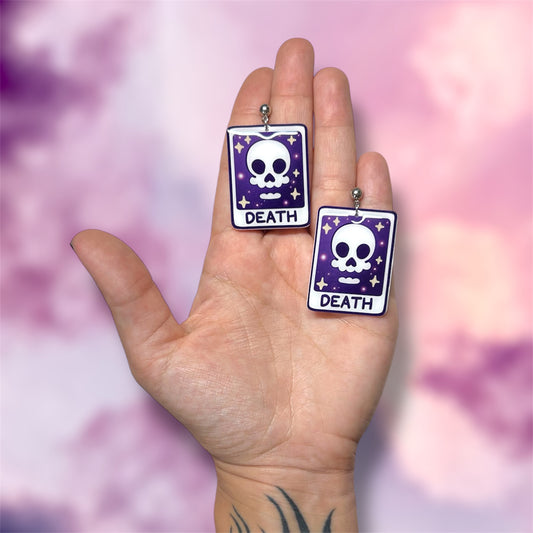 Hand holding sparkly skull Death tarot card witch resin dangle earrings on a blurry pink, purple, and white background