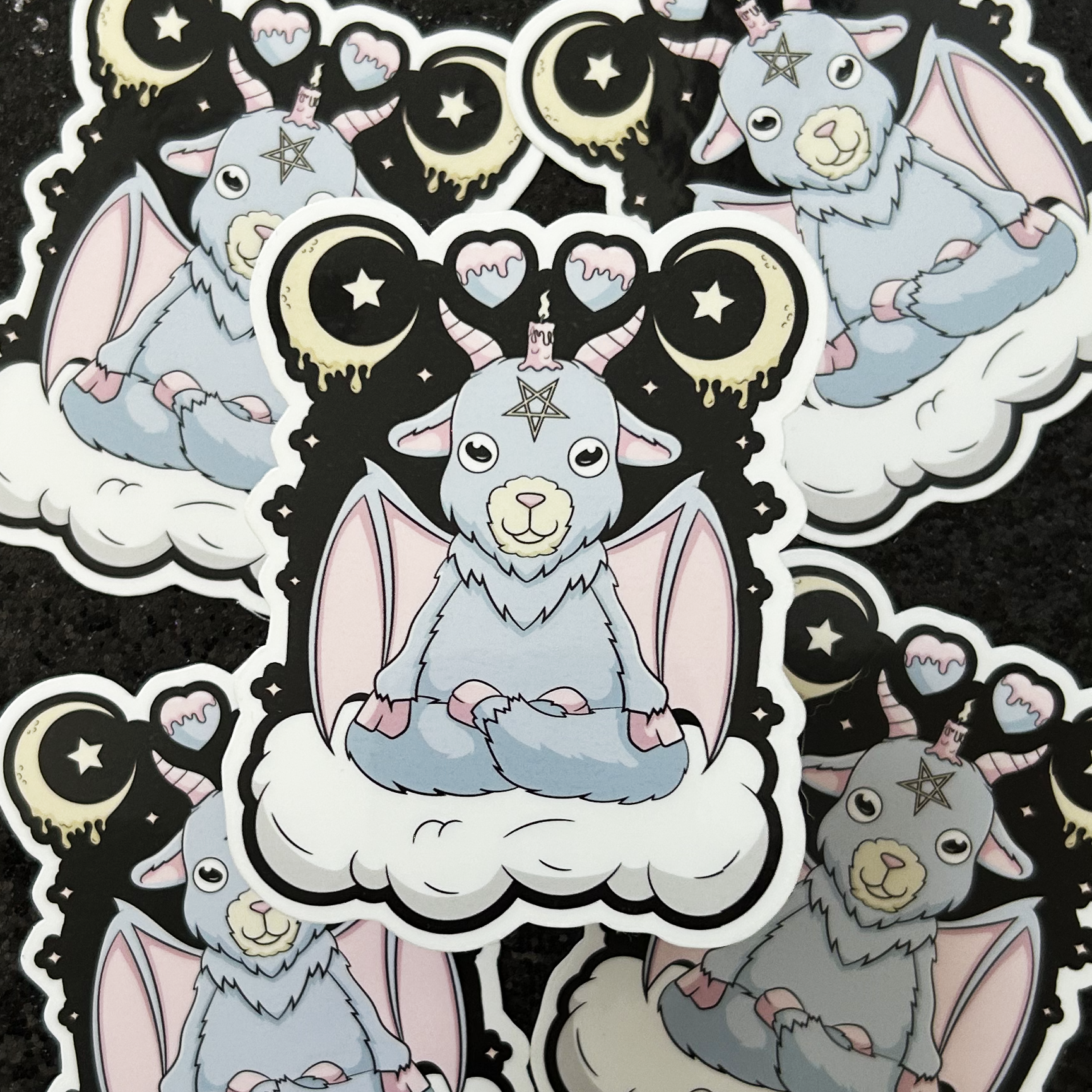 Stickers of a pastel-colored baphomet demon goat sitting on a cloud with celestial, witchy designs.