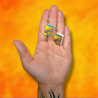 A hand holding a pair of Animorphs The Departure miniature book dangle earrings on a blurred orange and yellow background
