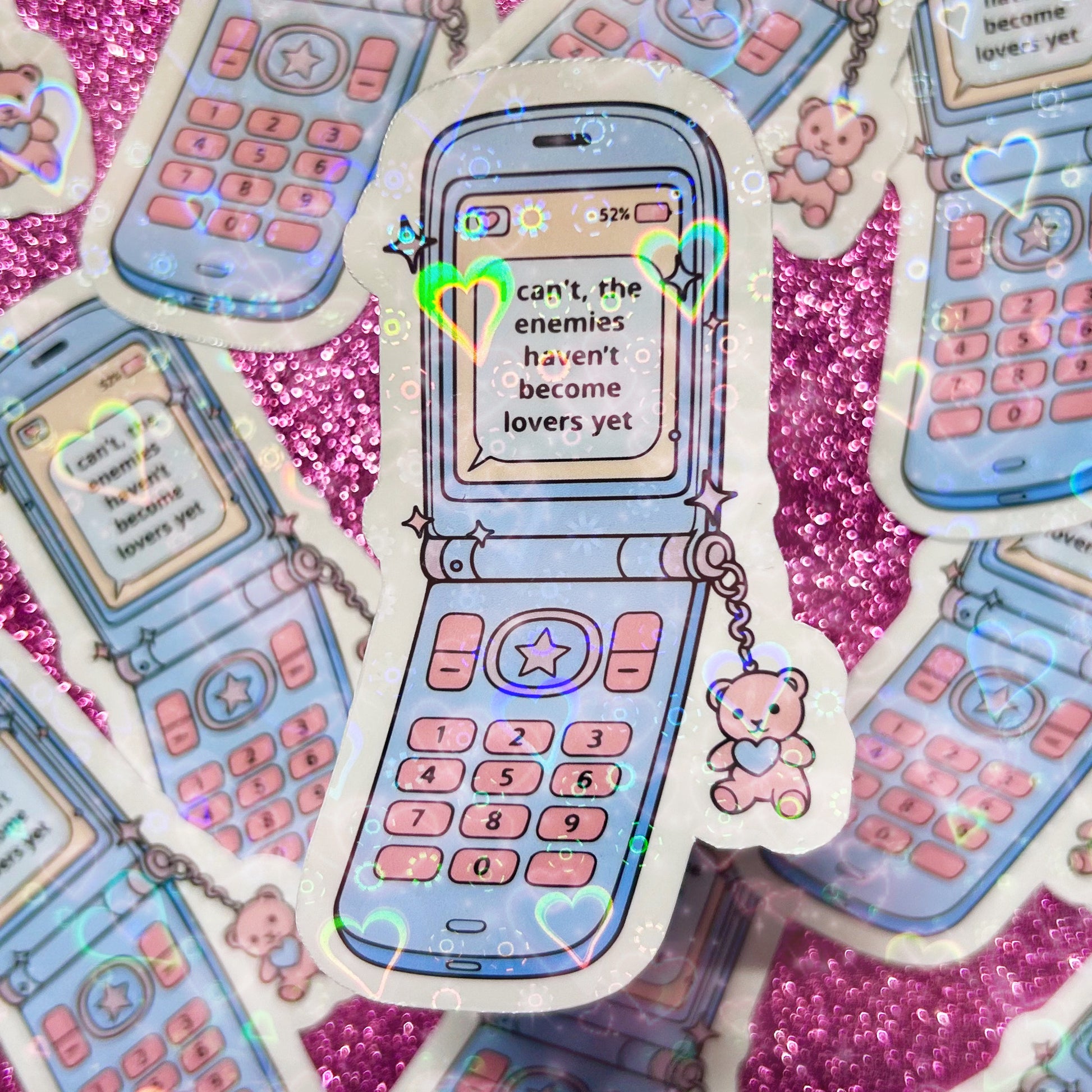 Sticker of a blue flip phone with a pink bear phone charm, the phone screen reads "I can't, the enemies haven't become lovers yet"
