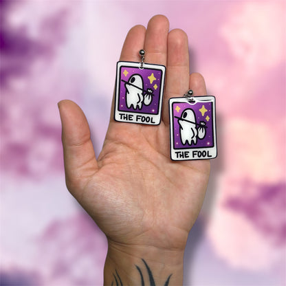 A hand holding a pair of The Fool tarot card witch resin dangle earrings on a blurred purple and pink background