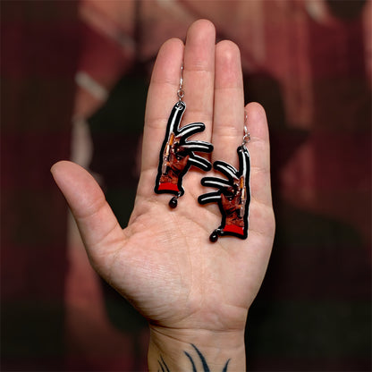 A hand holding a pair of Nightmare on Elm Street Freddy Krueger knife hand beaded resin statement earrings on a blurred red and black background