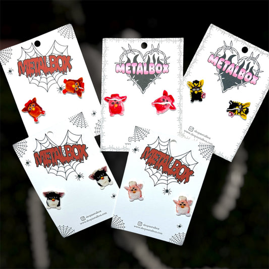 Five pairs of Furby resin stud earrings on a blurry background
