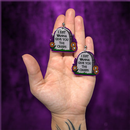 Hand holding spooky skull flower tombstone "I just wanna give you the creeps" resin statement earrings on a blurry purple and black background