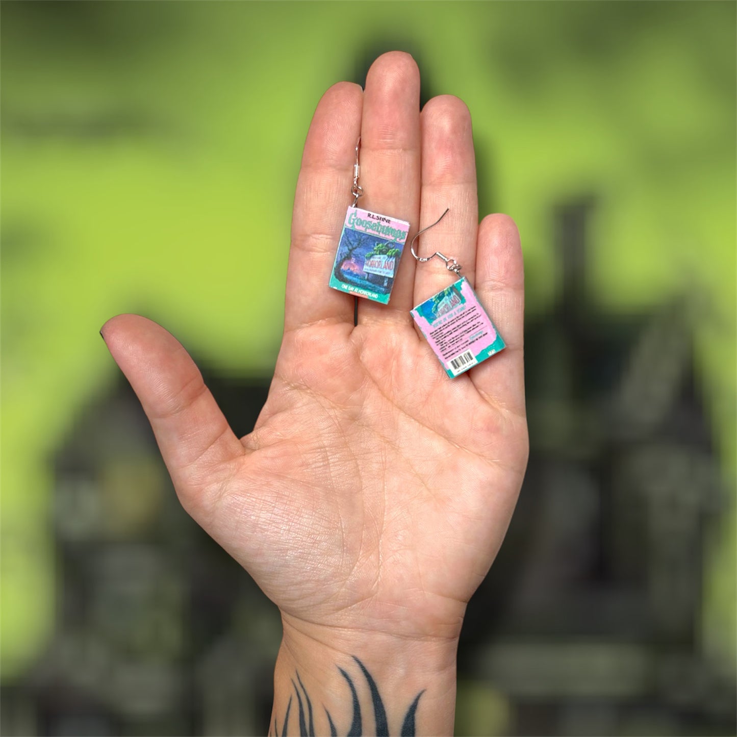 Hand holding a pair of Goosebumps nostalgic horror miniature book dangle earrings on a blurry green background