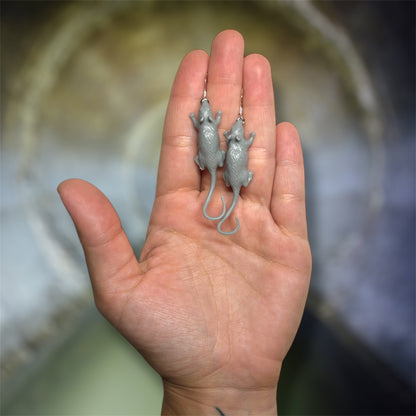 A hand holding a pair of gray rat dangle earrings on a blurred sewer background