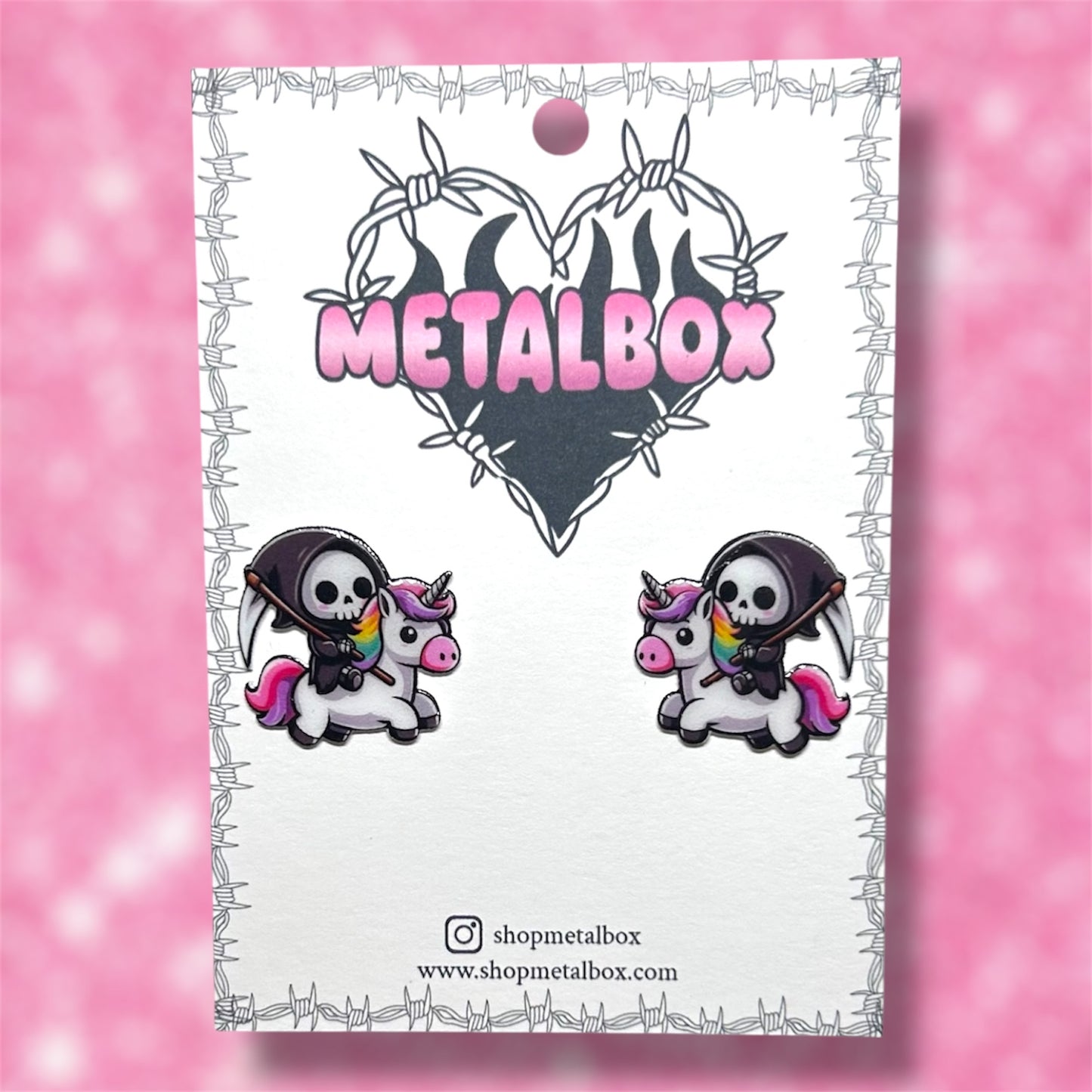 Grim Reaper holding a scythe riding a rainbow unicorn stud earrings on a Metalbox branded backing card on a blurry pink background