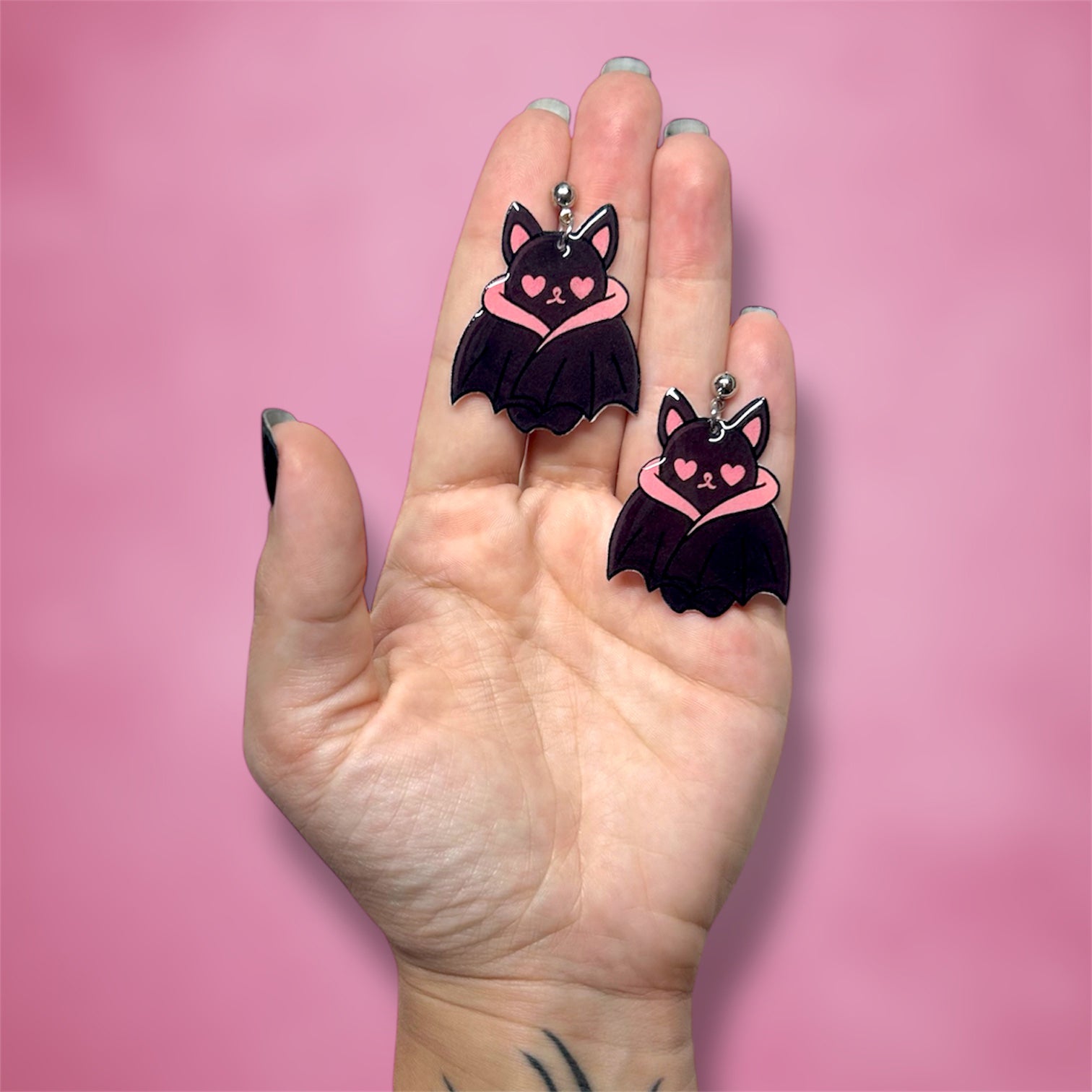 Hand holding a pair of bat earrings with pink heart eyes on a pink background