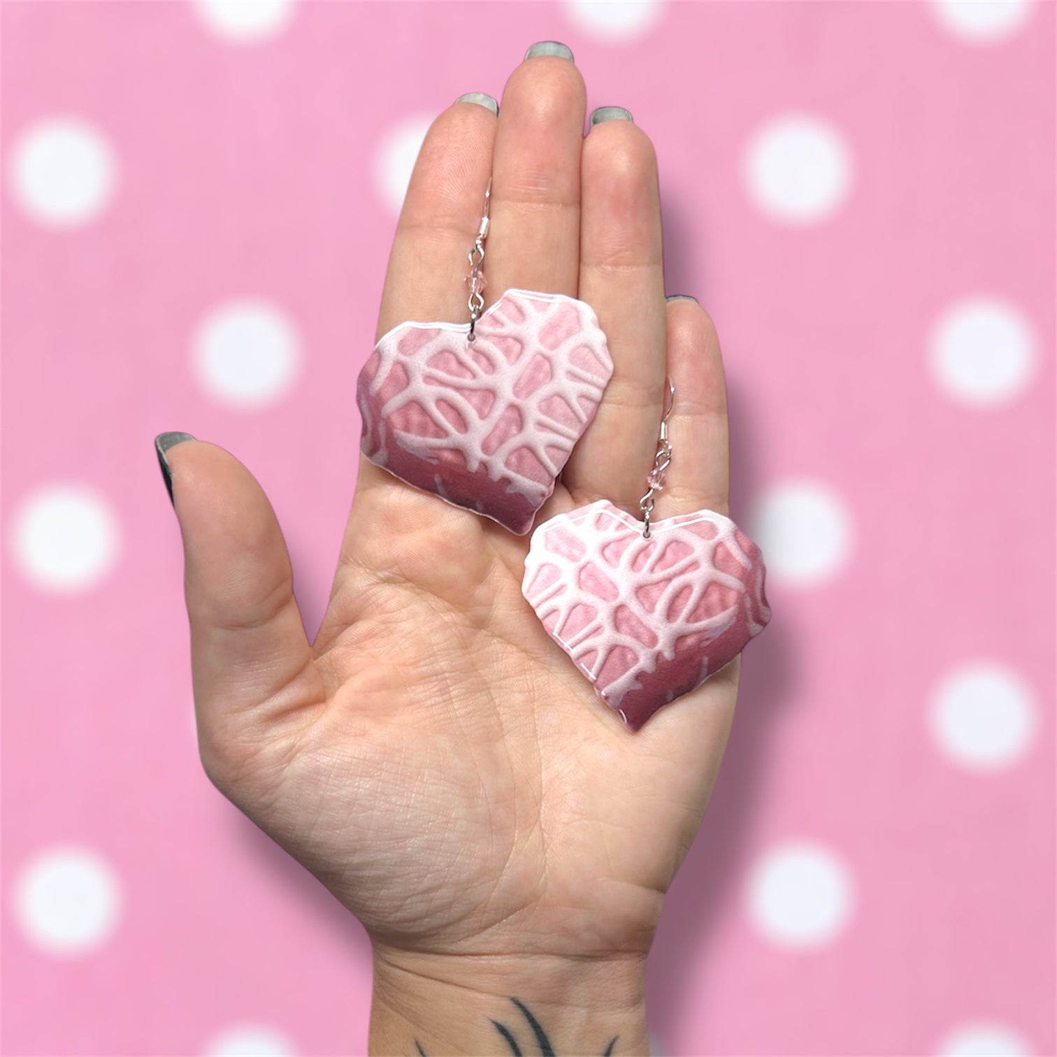 Hand holding a pair of Little Debbie Valentine's Day heart snack cakes beaded resin dangle earrings on a blurred pink background