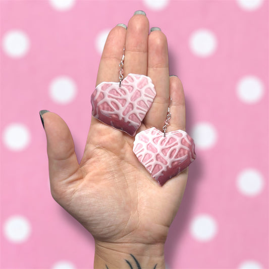 Hand holding a pair of Little Debbie Valentine's Day heart snack cakes beaded resin dangle earrings on a blurred pink background