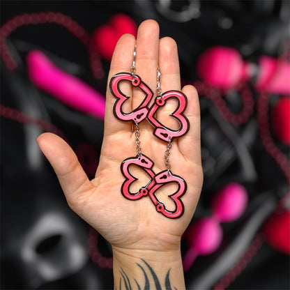 Hand holding a pair of heart-shaped pink and black handcuff dangle earrings on a blurred pink and black background