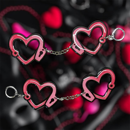 Heart-shaped pink and black handcuff dangle earrings on a blurred pink and black background
