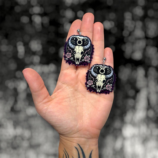 Hand holding a pair of Taurus zodiac sign skeleton floral bull skull dangle earrings on a blurry black and white background