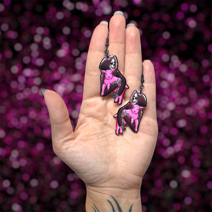 Hand holding a pair of earrings of a Grim Reaper skeleton holding a pink lamb on a blurred glittery pink background.