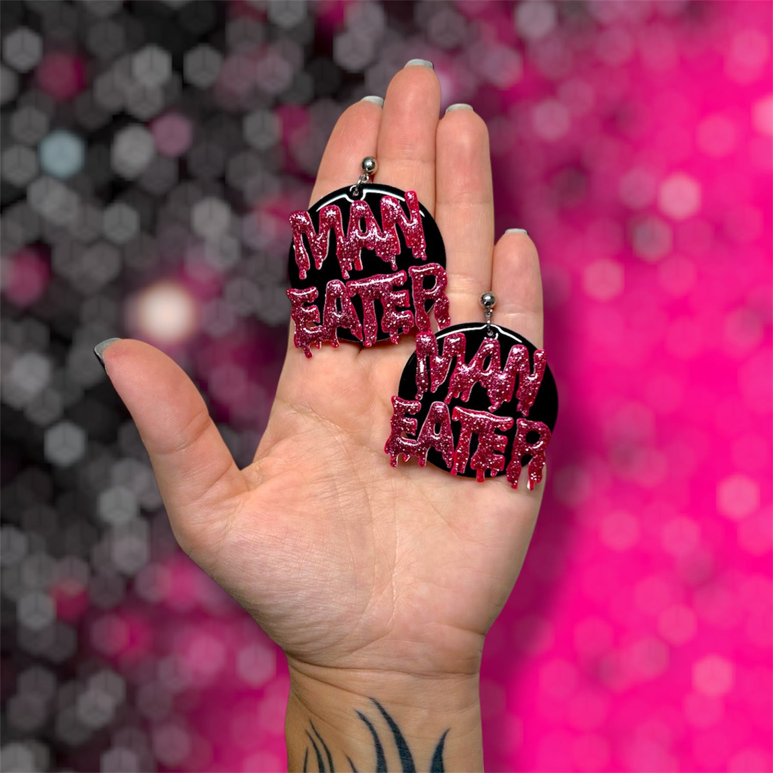 Hand holding a pair of Man Eater drippy glittery resin round dangle earrings on pink and black blurred background